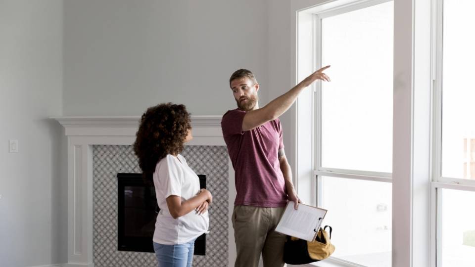 Home inspector points to something outside of a window while talking with pregnant homeowner