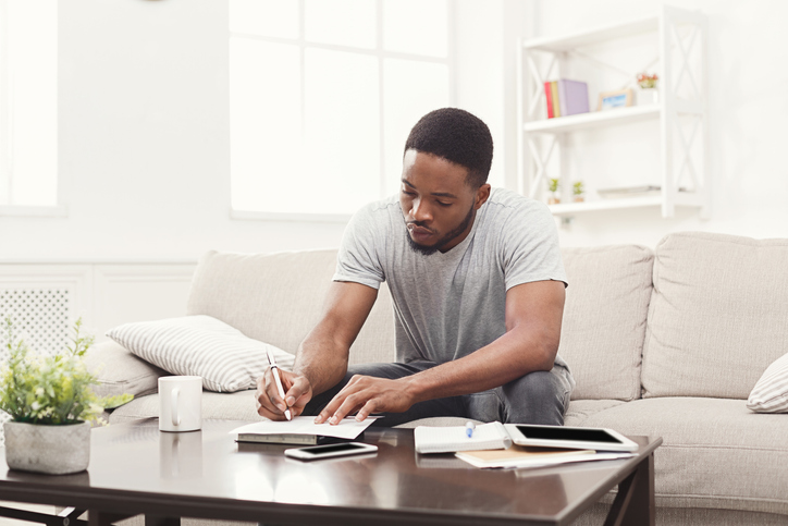 Young man sitting on couch at home, concentrating while taking notes
