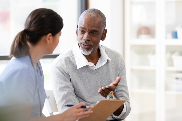 Man gestures while speaking to a female nurse. The nurse holds a clipboard and is documenting information about the patient.