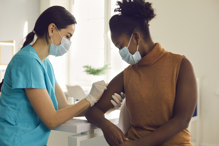 Nurse vaccinating young female patient inside a clinic, wearing protective face masks