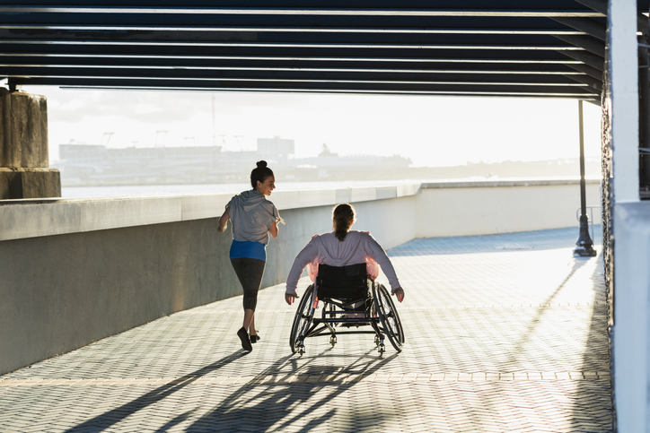 Rear view of two friends exercising along a city waterfront. The young woman in the wheelchair has spina bifida.