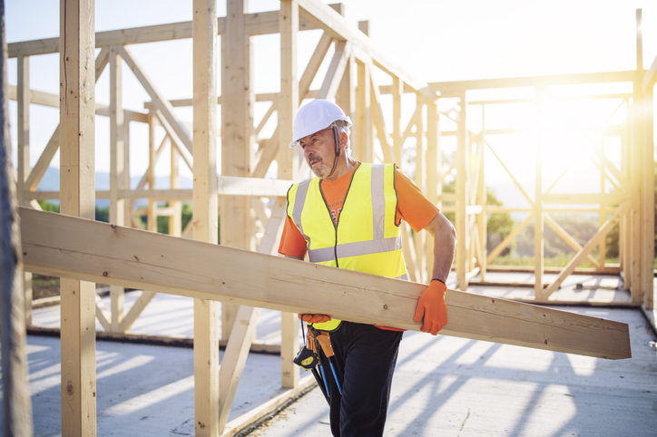 Side view of builder holding wooden beam, working on unfinished wooden house at sunset